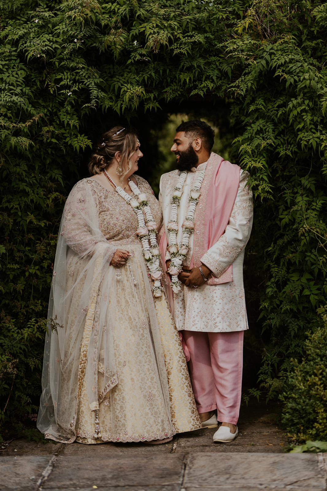 Sherni London bride and groom in traditional Indian lehenga and sherwani in cream gold and pink standing in garden setting with floral garland haar