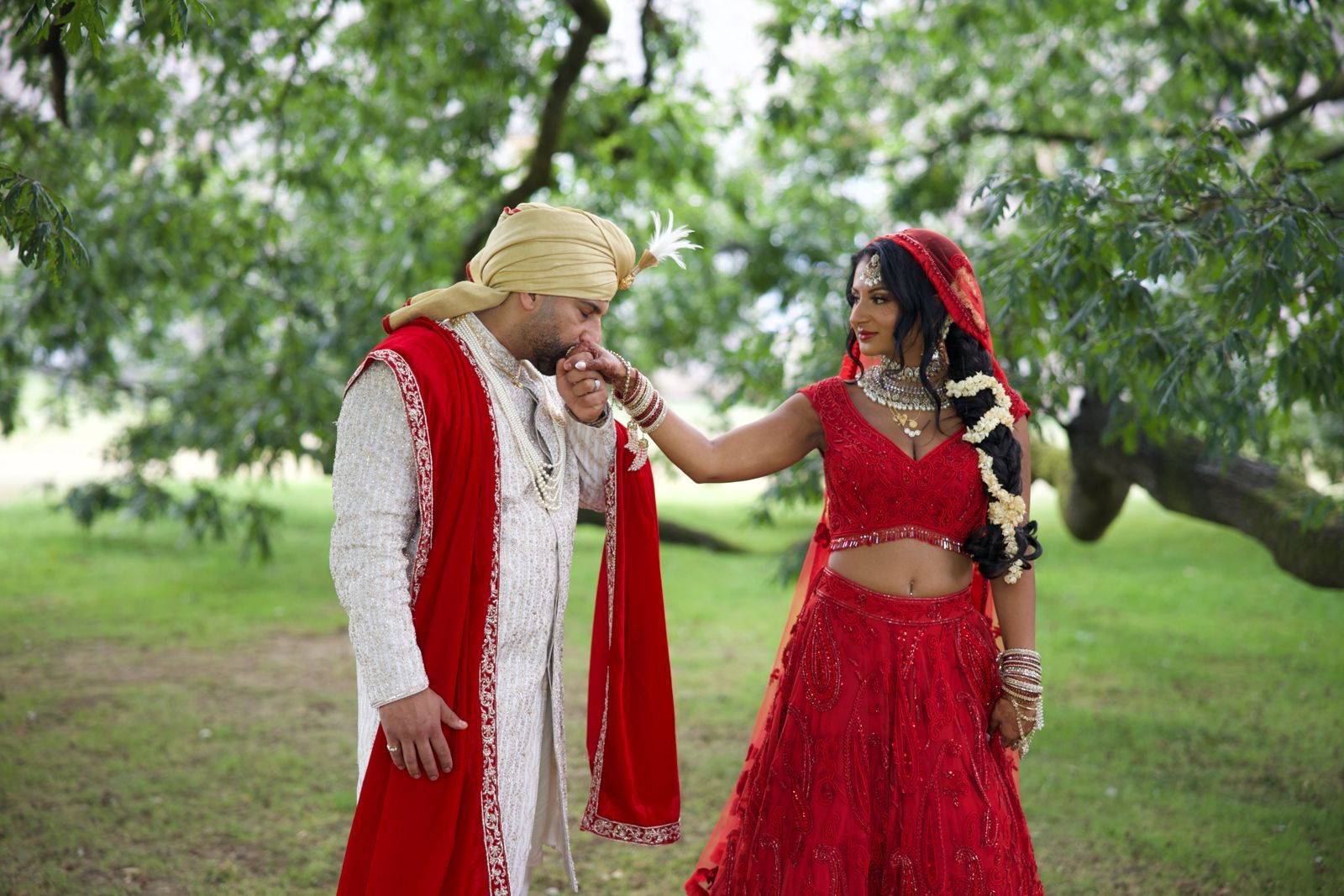 Bride and groom in sherwani and red lehenga in nature kissing hand photoshoot with flowers