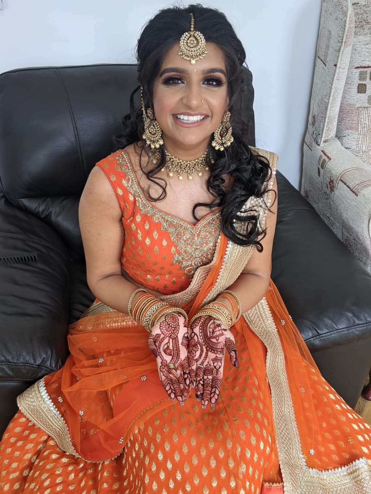 Sherni London bride woman in traditional Indian orange and gold lehenga with jewellery and henna, sitting on a black couch.