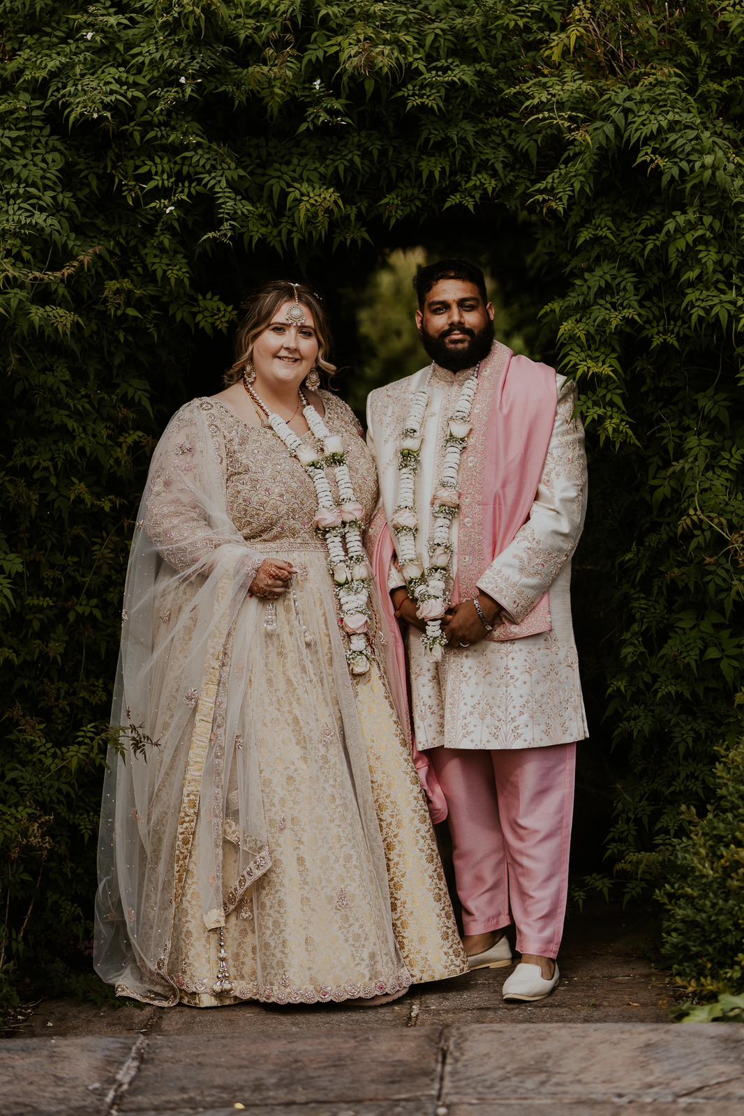 Sherni London bride and groom in traditional Indian lehenga and sherwani in cream gold and pink standing in greenery with floral garland haar around their necks