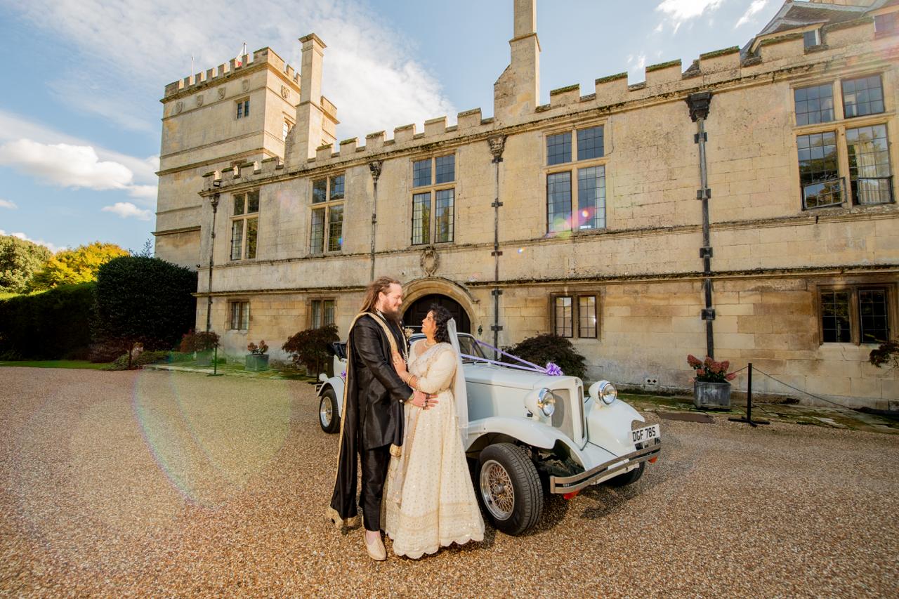 Sherni London bride and groom in fusion church wedding ceremony wearing white chikankari lucknowi lehenga and black asymmetric sherwani with gold buttons and trim standing in front of vintage car
