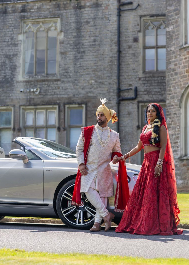 Bride and groom in sherwani and red lehenga traditional attire standing next to a silver convertible car in front of a castle.