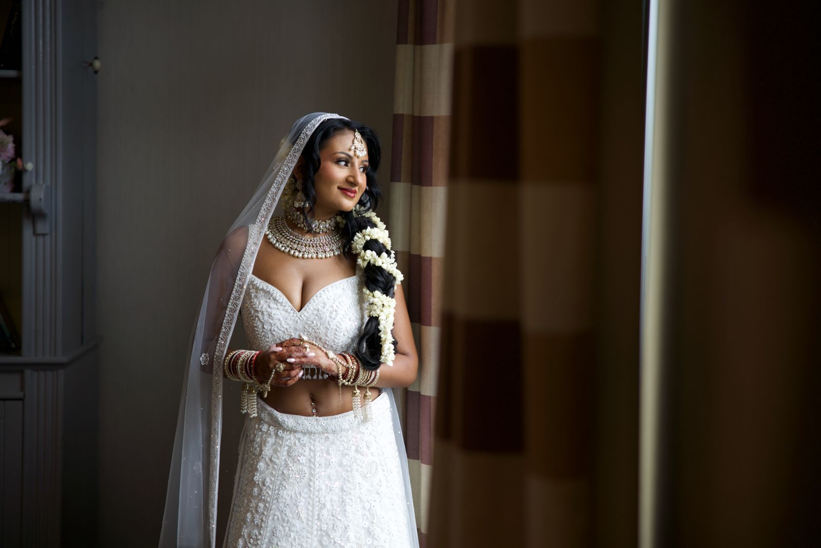 South Asian bride in white lehenga with hand embroidery all over and white net dupatta veil on her head with flowers in her hair and bangles 