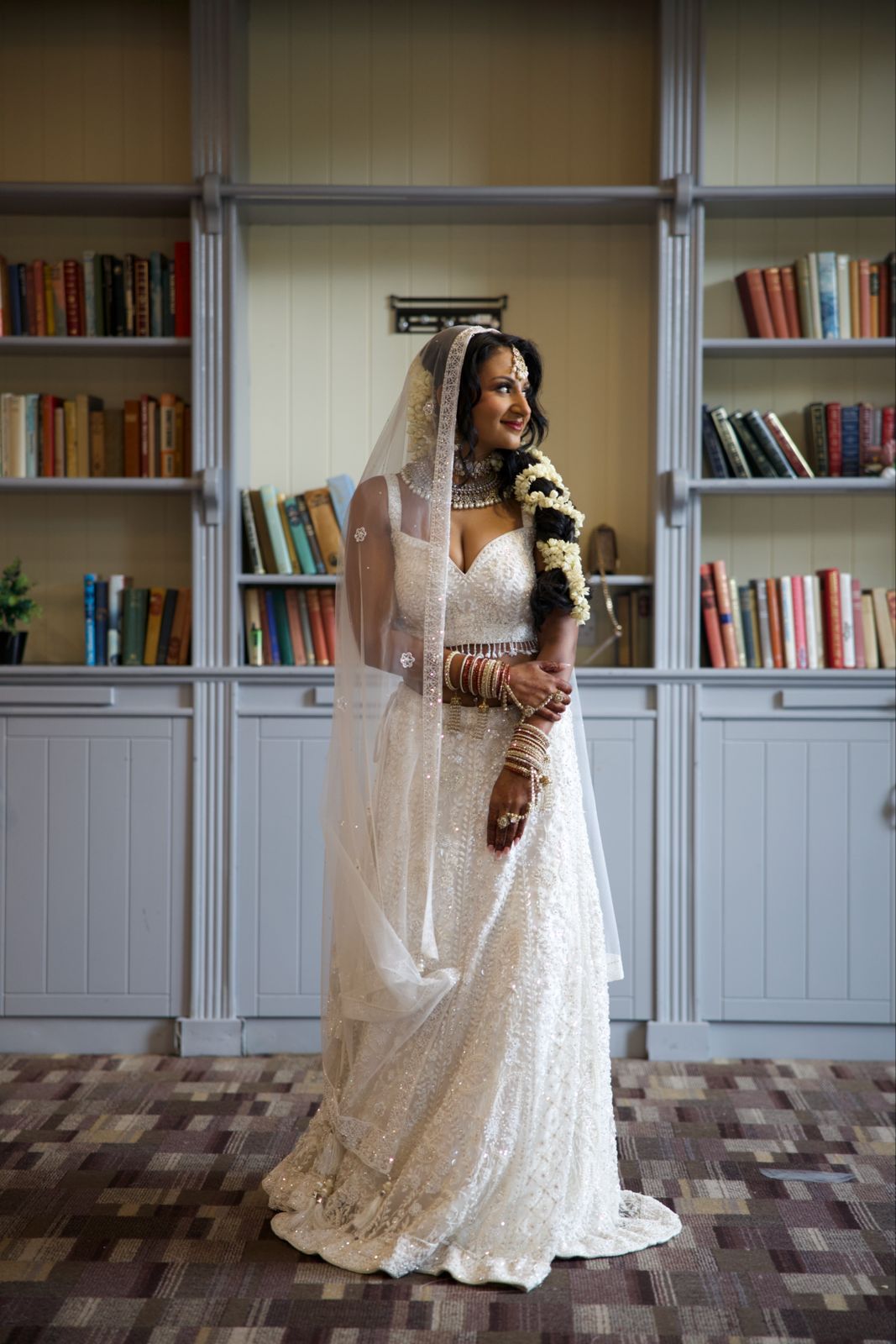 South Asian bride in white lehenga with hand embroidery all over and white net dupatta veil on her head with flowers in her hair and bangles standing in front of wall of books library