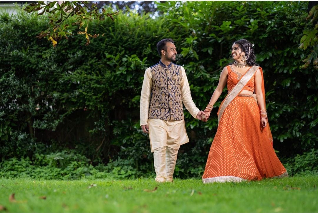 Sherni London couple holding hands in orange Indian bridal lehenga against a green hedge background