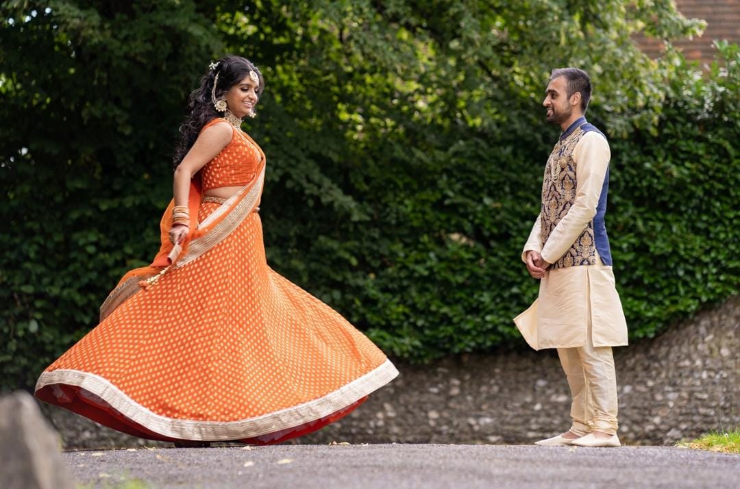 Woman in traditional orange lehenga for her engagement sagai dancing outdoors with a man in formal attire standing nearby.